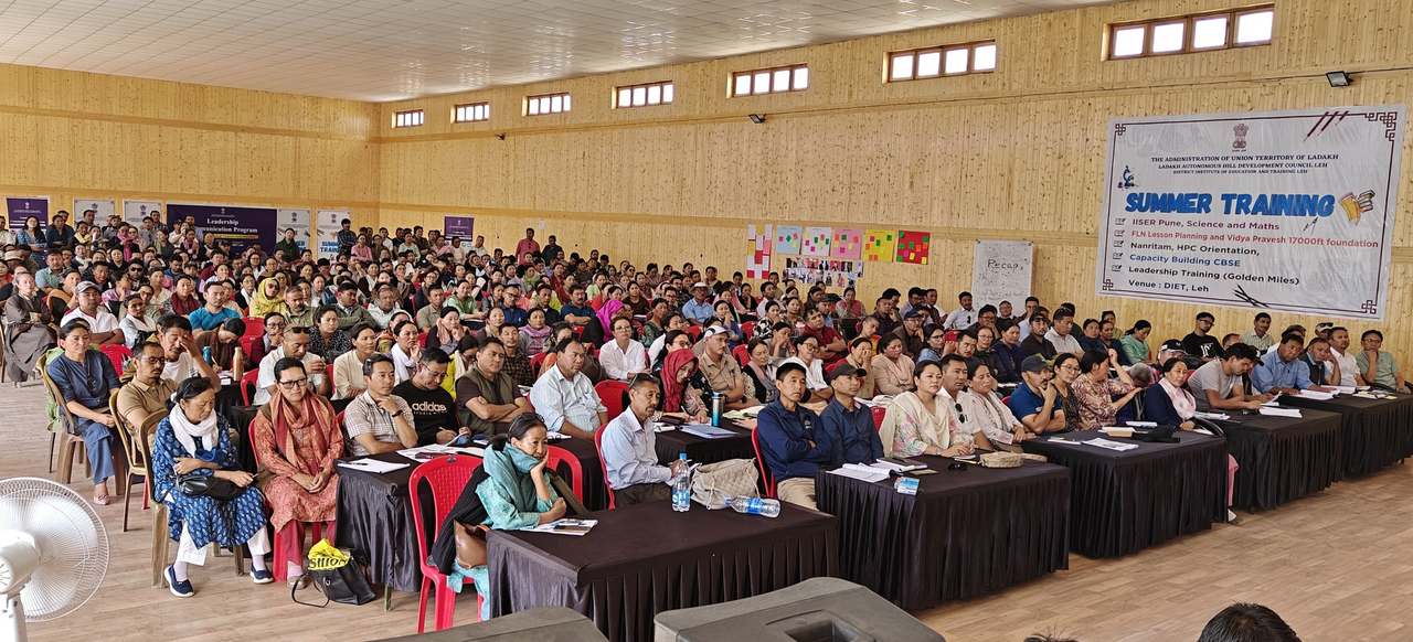 Rows of teachers seated at tables in a wood-panelled hall during DIET Leh's Summer Training, with the training banner on the wall